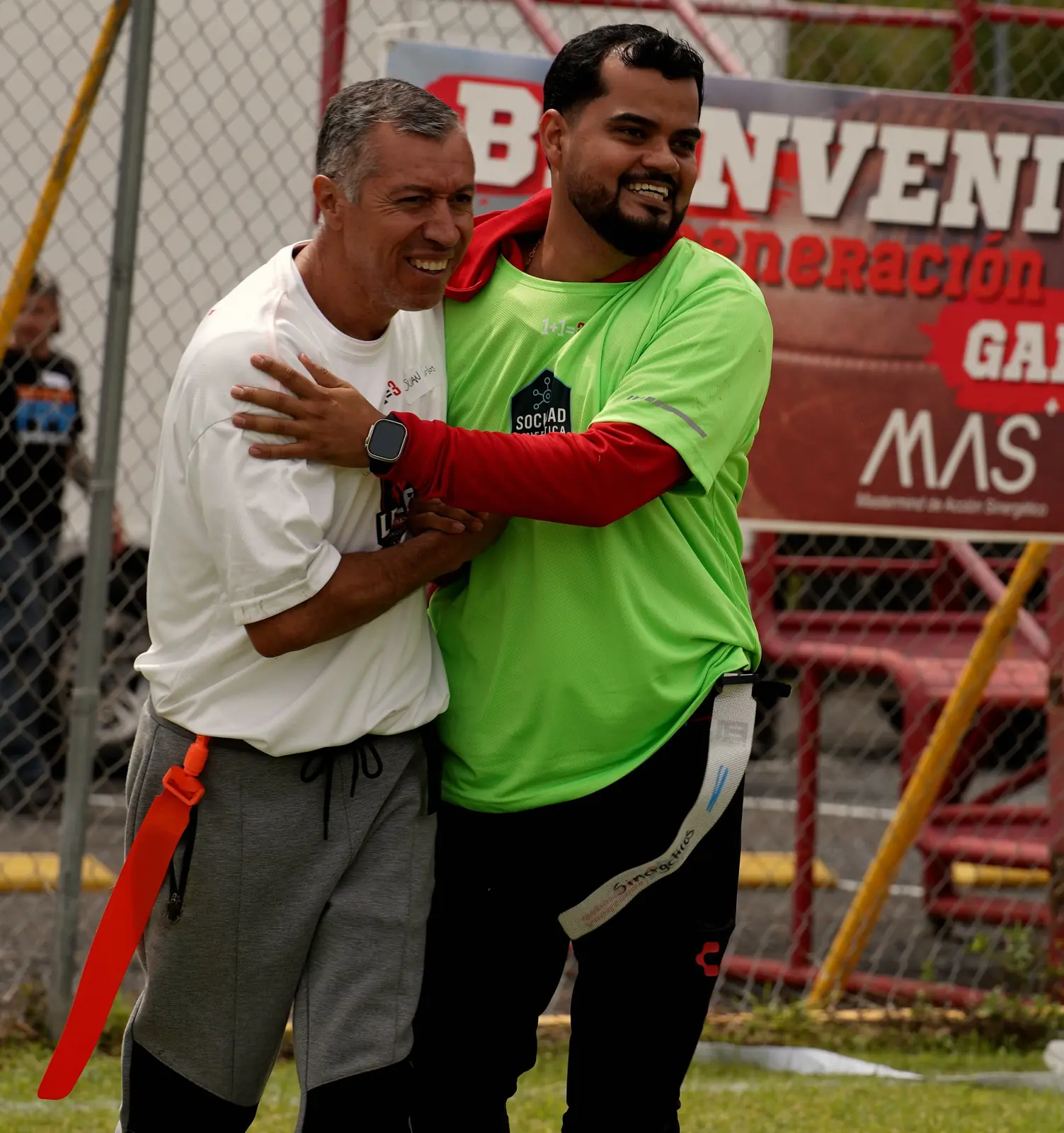 Dos integrantes se abrazan riendo frente a la lona de bienvenida del día del torneo del MAS