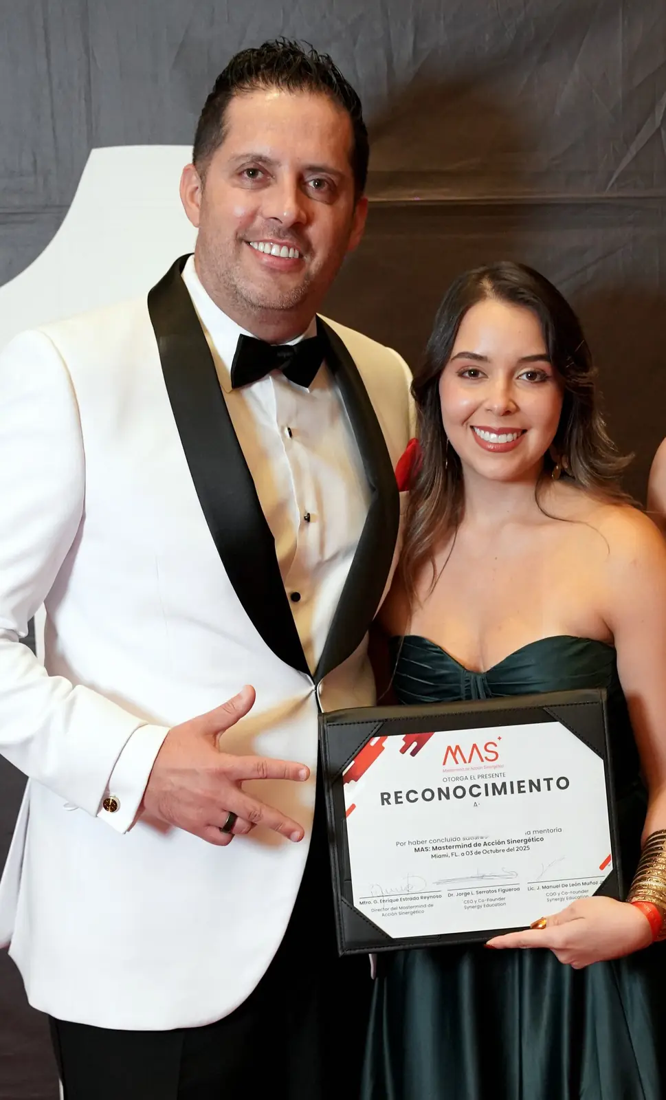Jorge Serratos, de smoking blanco, señala orgulloso a una graduada del MAS con su reconocimiento