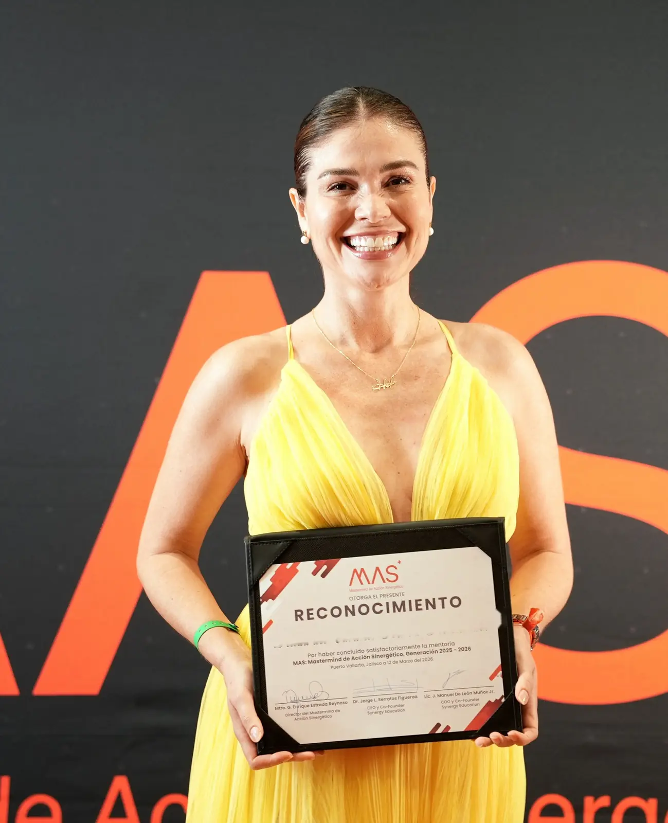 Una graduada del MAS en vestido amarillo sostiene su reconocimiento frente al logotipo del MAS